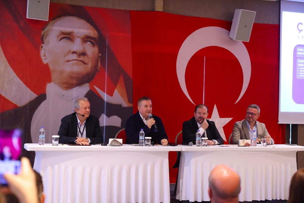 Panel of four men seated at a long white table on a stage, microphones and water bottles in front, with a large Turkish flag backdrop and Atatürk portrait behind them.
