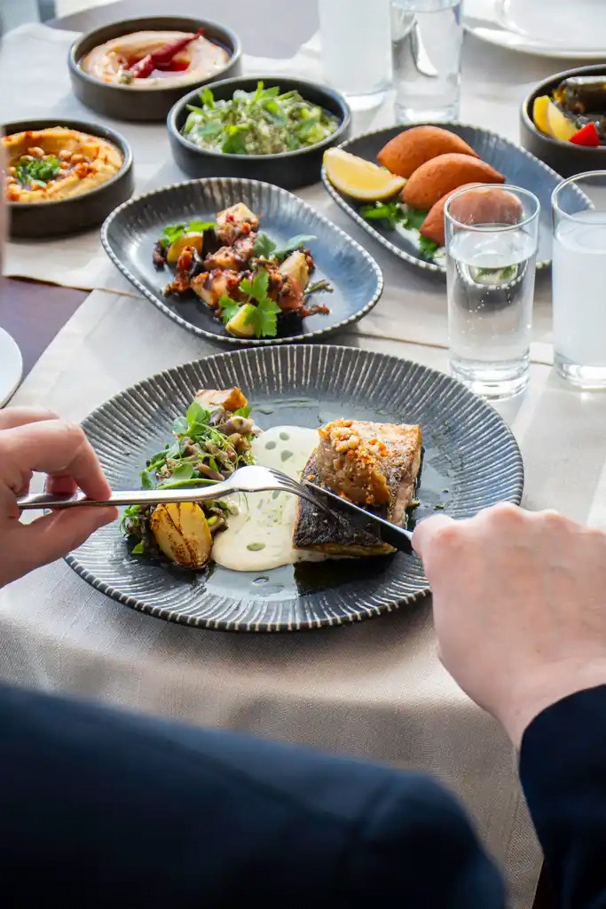 A person cuts a seared fish fillet on a sauced plate with greens, surrounded by multiple side dishes on a dining table.