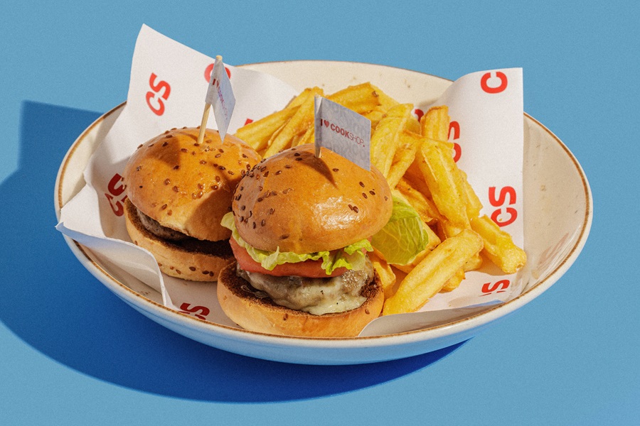 Two cheeseburgers with lettuce and tomato and a side of french fries in a paper-lined white bowl on a bright blue surface.