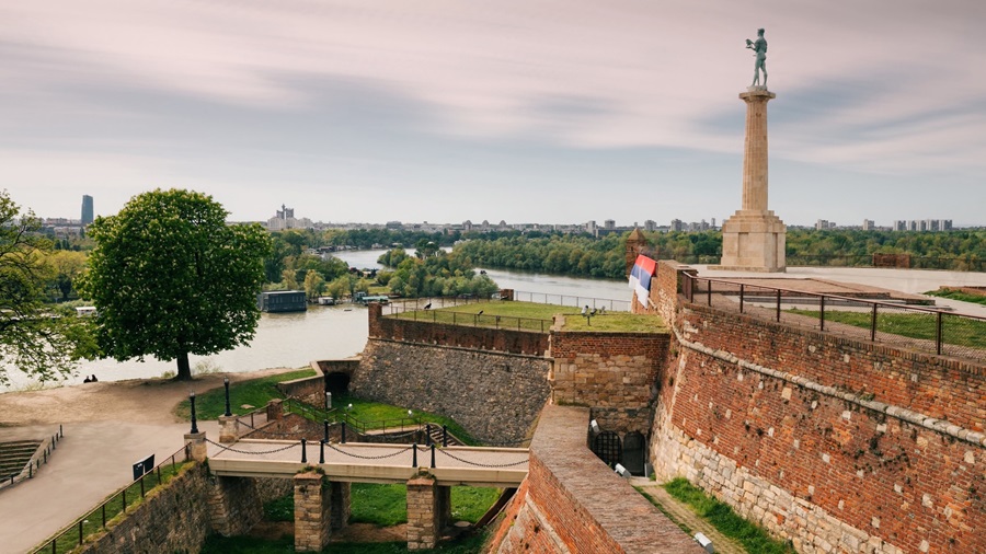 Brick fortress along a river, with tree-lined banks, ramparts, and a statue on a tall column at the edge nearby a flag. (Informative)