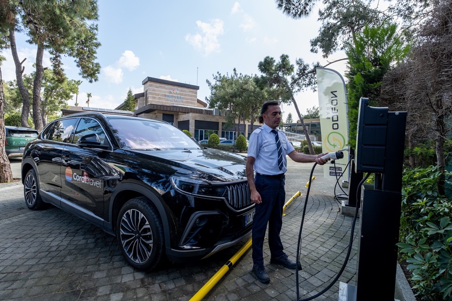 Man in uniform plugs in an electric car at an outdoor charging station beside a corporate building with trees around.