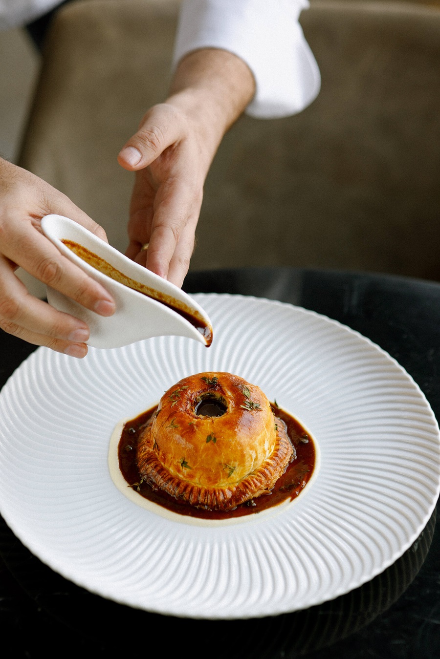 Chef pours dark sauce over a circular puff pastry dish plated on a white textured plate.
