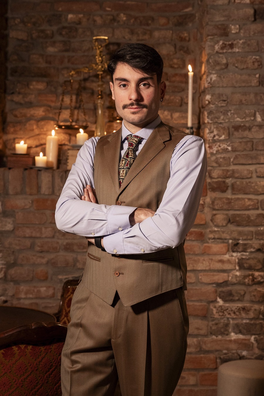 Portrait of a man with a mustache in a tan vest and suit, arms crossed, in a warm candlelit brick room.