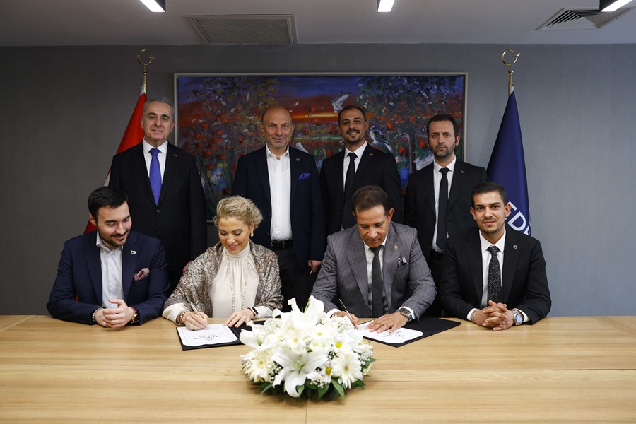 Formal signing ceremony: two people sign documents at a table while six colleagues stand behind, with a bouquet and flags in the background.