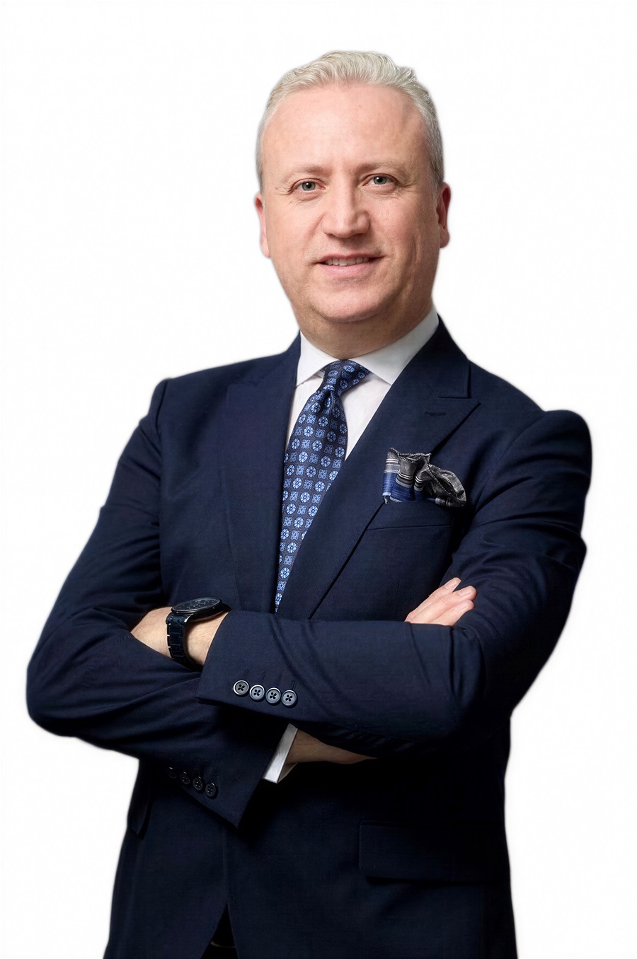 Professional portrait of a middle-aged man with silver hair in a navy suit, patterned blue tie, and pocket square, standing with arms crossed against a white background.