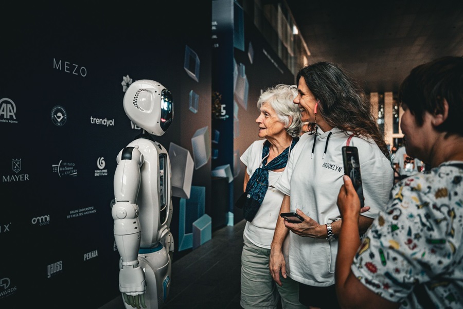 A white humanoid robot standing beside two casually dressed women at an event, as they interact and smile near a dark wall with logos.