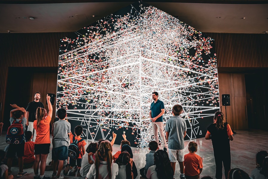 Audience of children watching a tech demonstration on stage with a large, illuminated geometric network sculpture in the background.