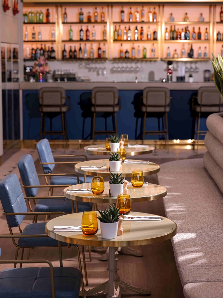 Chic bar seating with round gold tables, blue chairs, orange glasses, and small potted succulents along a backlit liquor shelves backdrop.