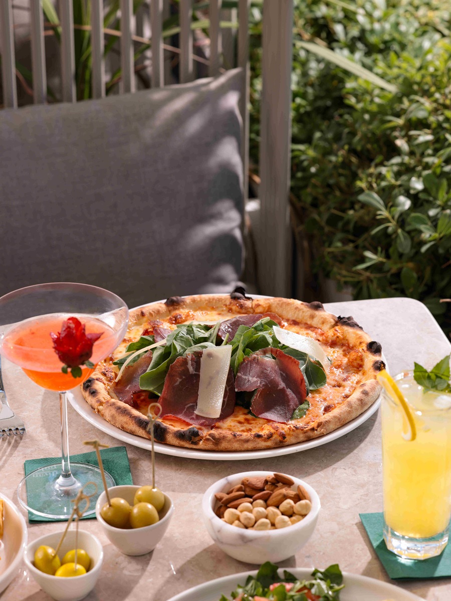 Outdoor dining scene with a wood-fired pizza topped with greens and shaved parmesan on a plate, surrounded by olives, nuts, and a yellow cocktail on a sunny table
