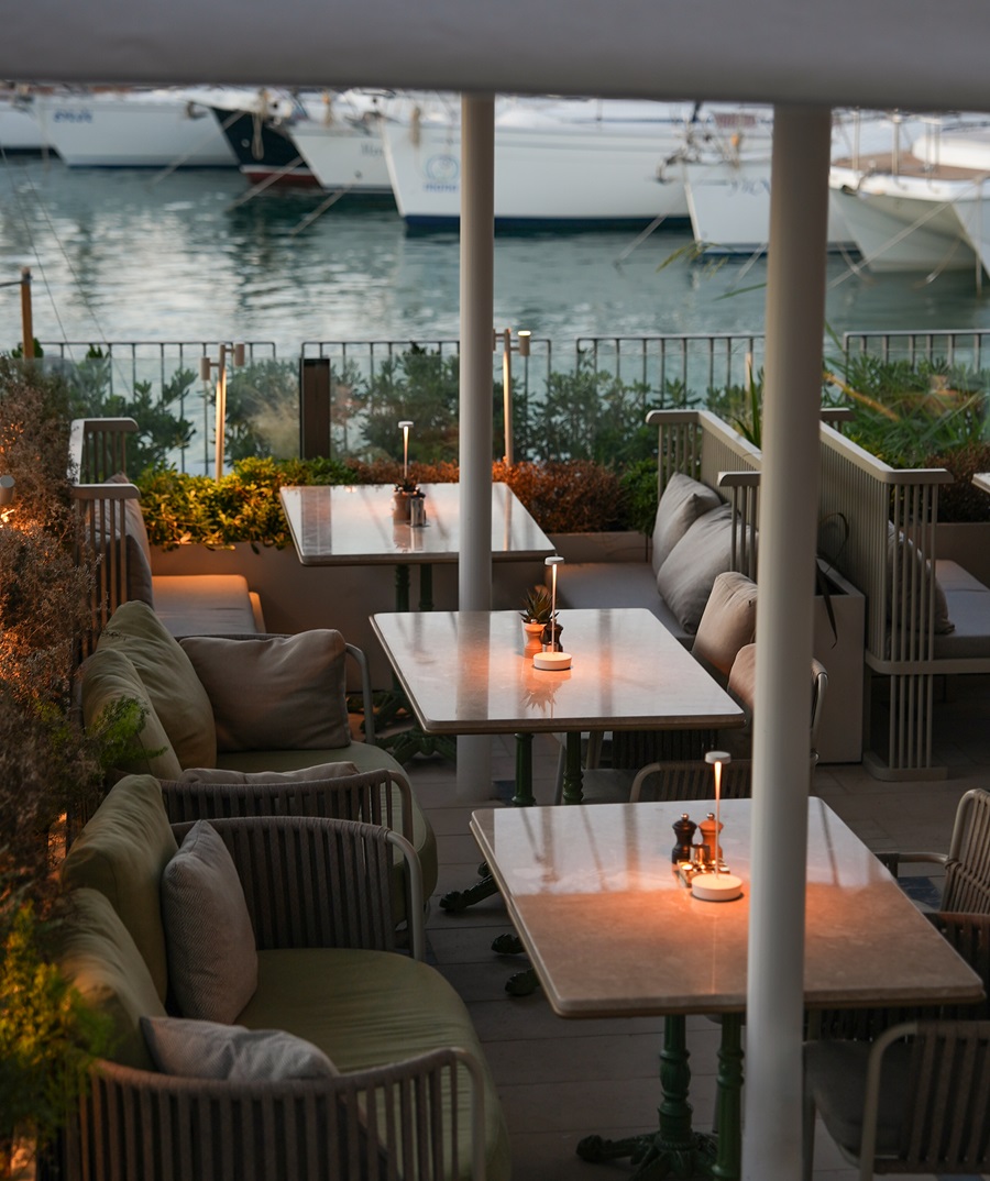 Cozy outdoor restaurant terrace by the marina at dusk with candlelit tables and cushioned seating by the water.