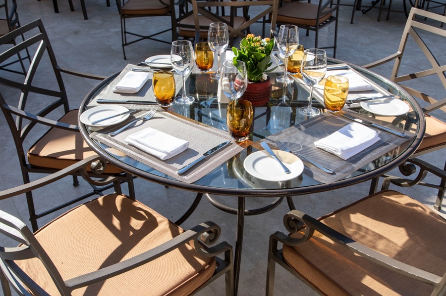 Round glass outdoor dining table set for four with napkins, plates, silverware, amber glasses, and a small flower centerpiece.