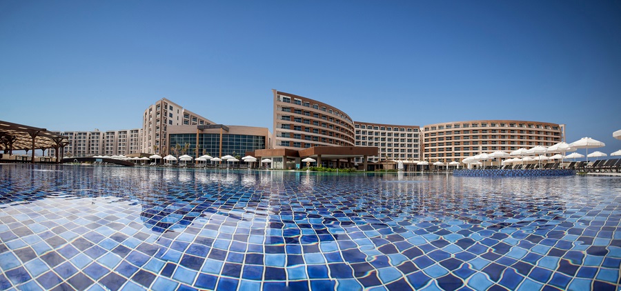Resort hotel with curved wings surrounding a large blue tiled pool and white lounge umbrellas under a clear sky.