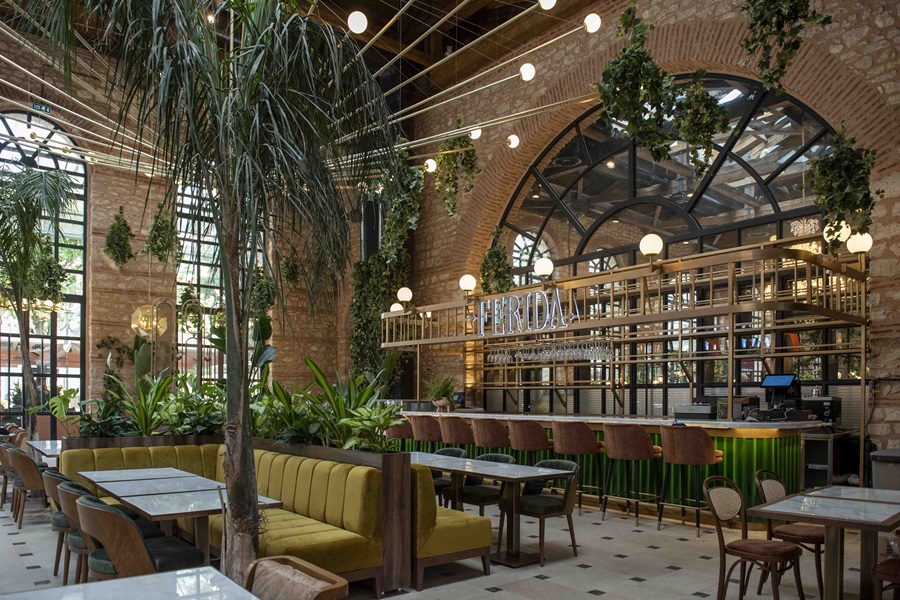 Bright, plant-filled cafe interior with brick arches, tall windows, yellow banquette seating, and a long green-bar counter behind glass shelves.