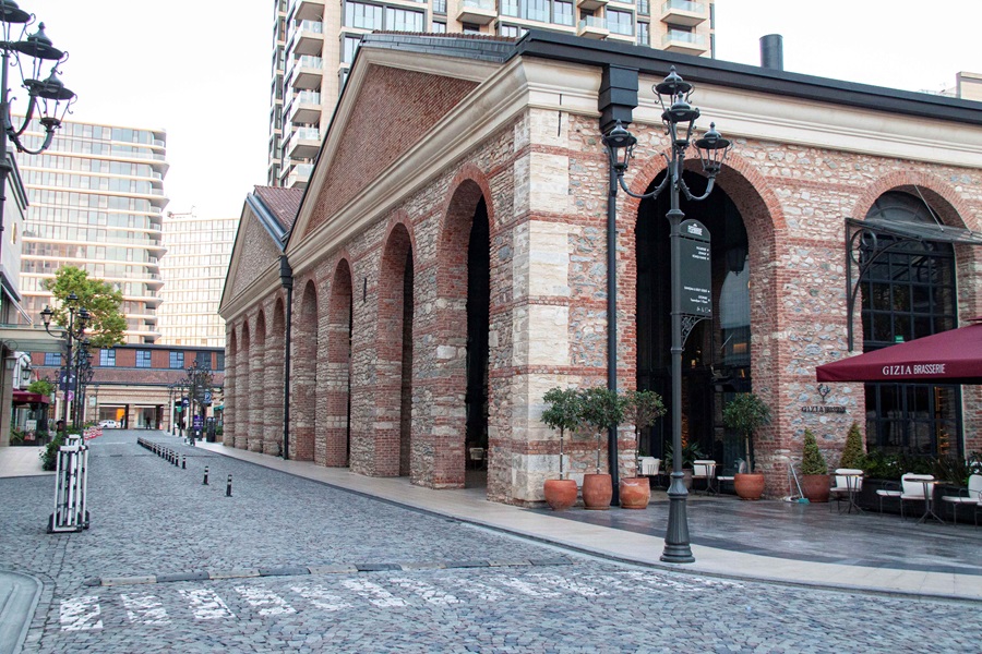 Brick arched arcade along a cobblestone street, with outdoor café seating and potted plants; modern buildings loom in the background.