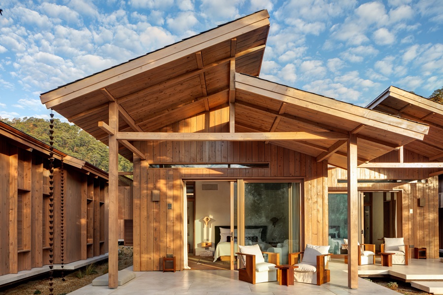 Wooden resort bungalow with a large overhanging roof, outdoor seating, and a bedroom visible through glass doors.