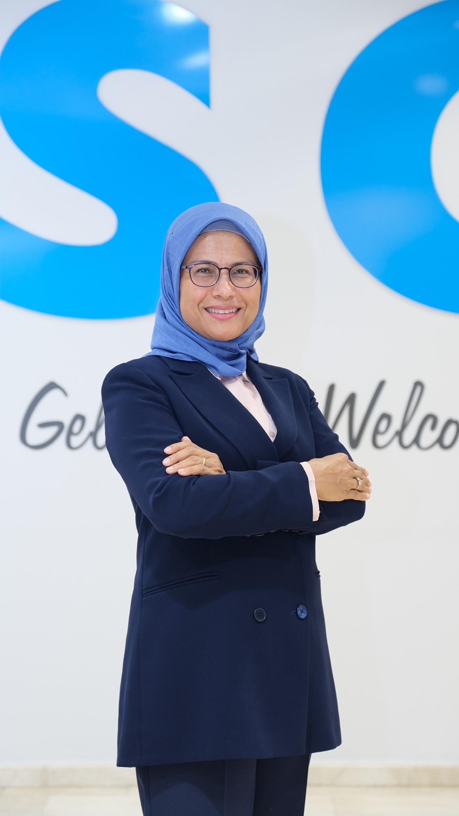Professional woman wearing a blue hijab and navy suit with arms crossed, standing in front of a welcome sign and blue logo.