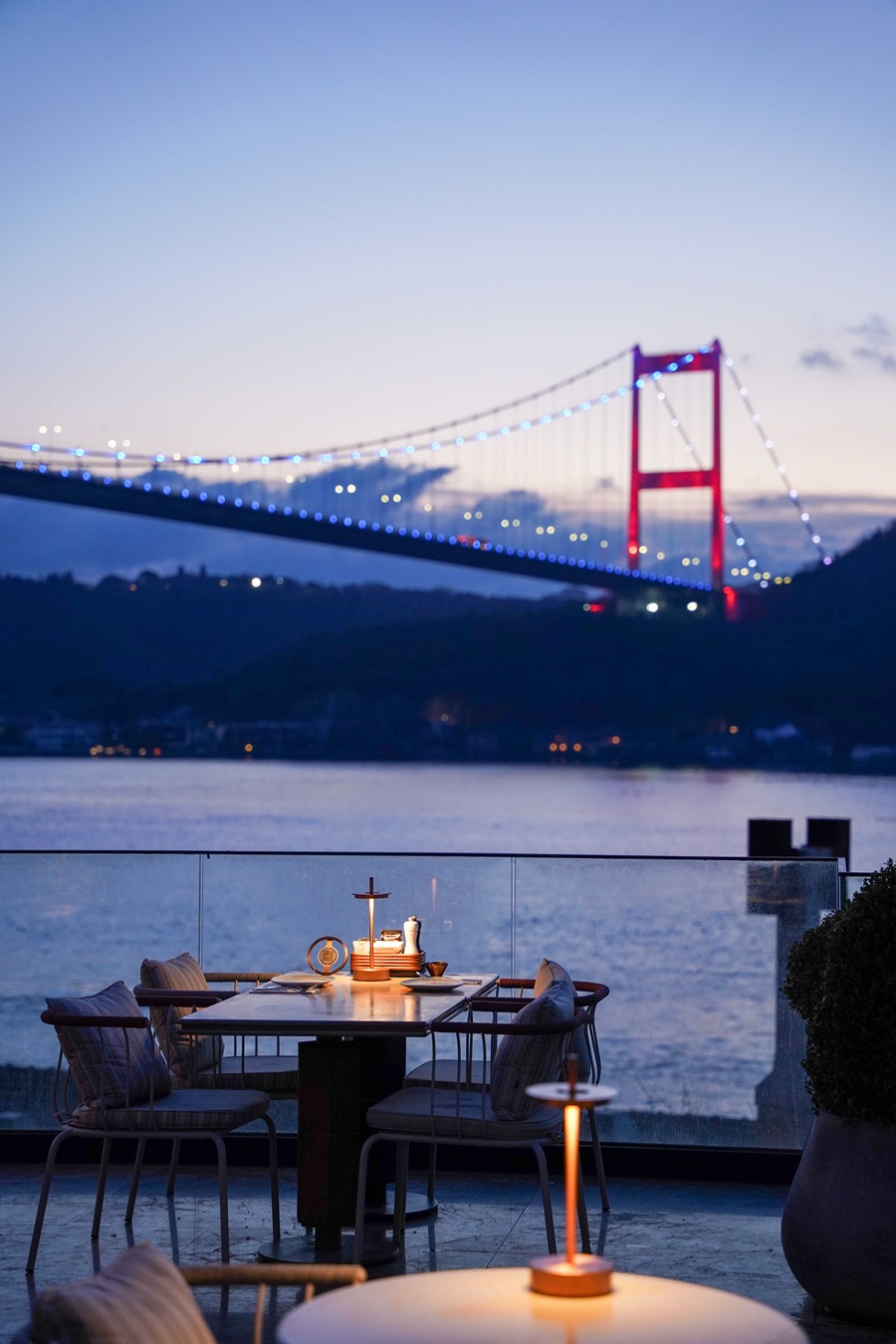 Outdoor dining table by the water with candles, overlooking a blue-lit suspension bridge at dusk.