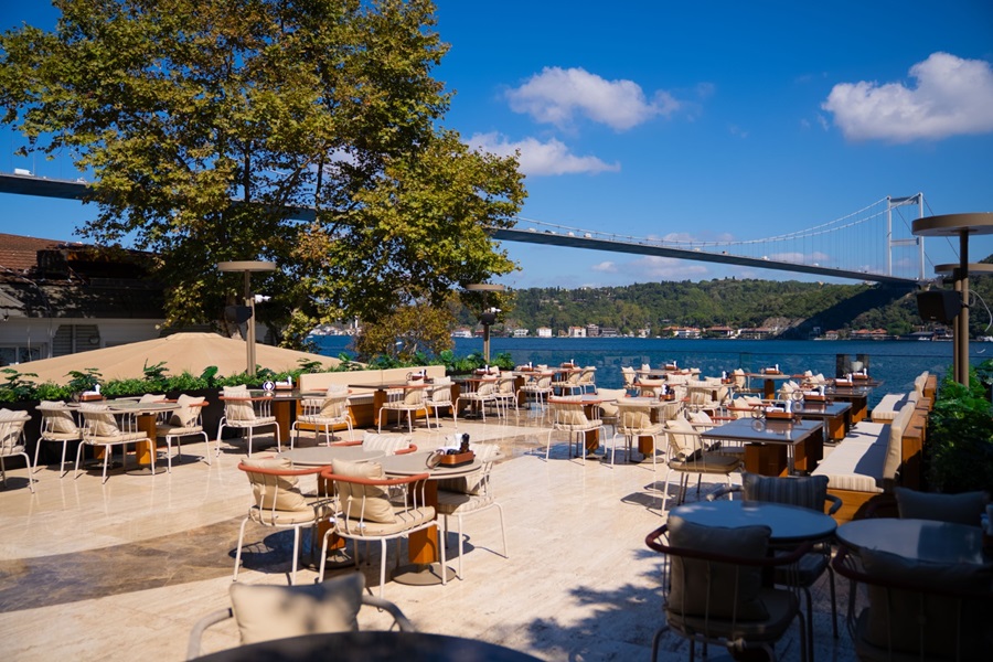 Outdoor restaurant terrace by the water with many beige tables and chairs, a large tree, and a suspension bridge in the background on a sunny day.