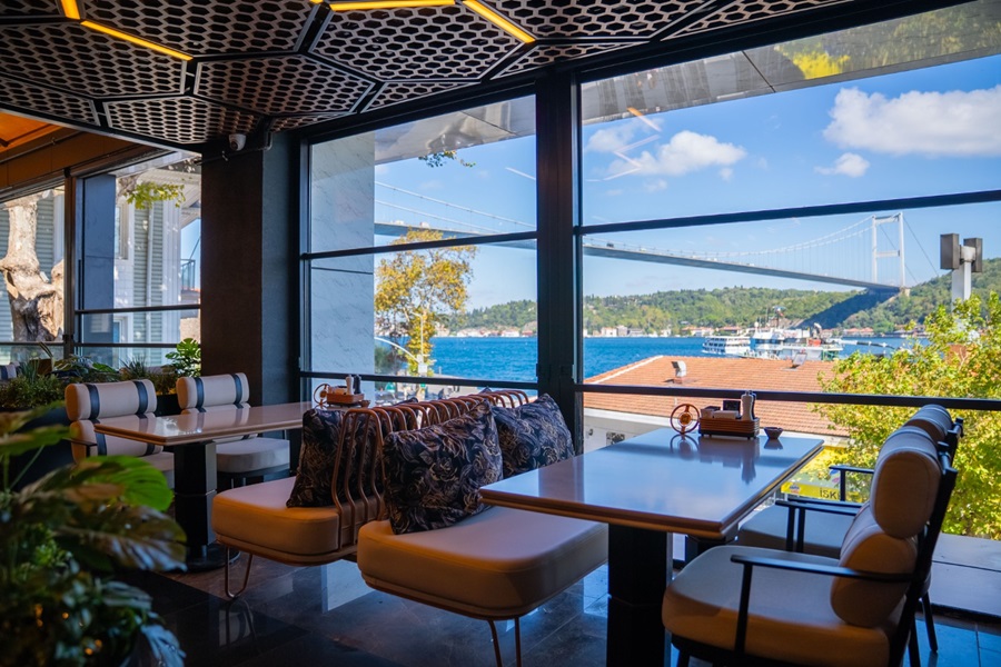 Cafe interior with cream leather seating and a long table, large windows show blue water and a distant bridge landscape.