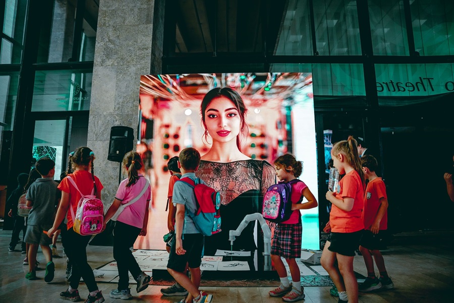 Children with backpacks gather around a large illuminated screen showing a portrait of a woman in a mall,