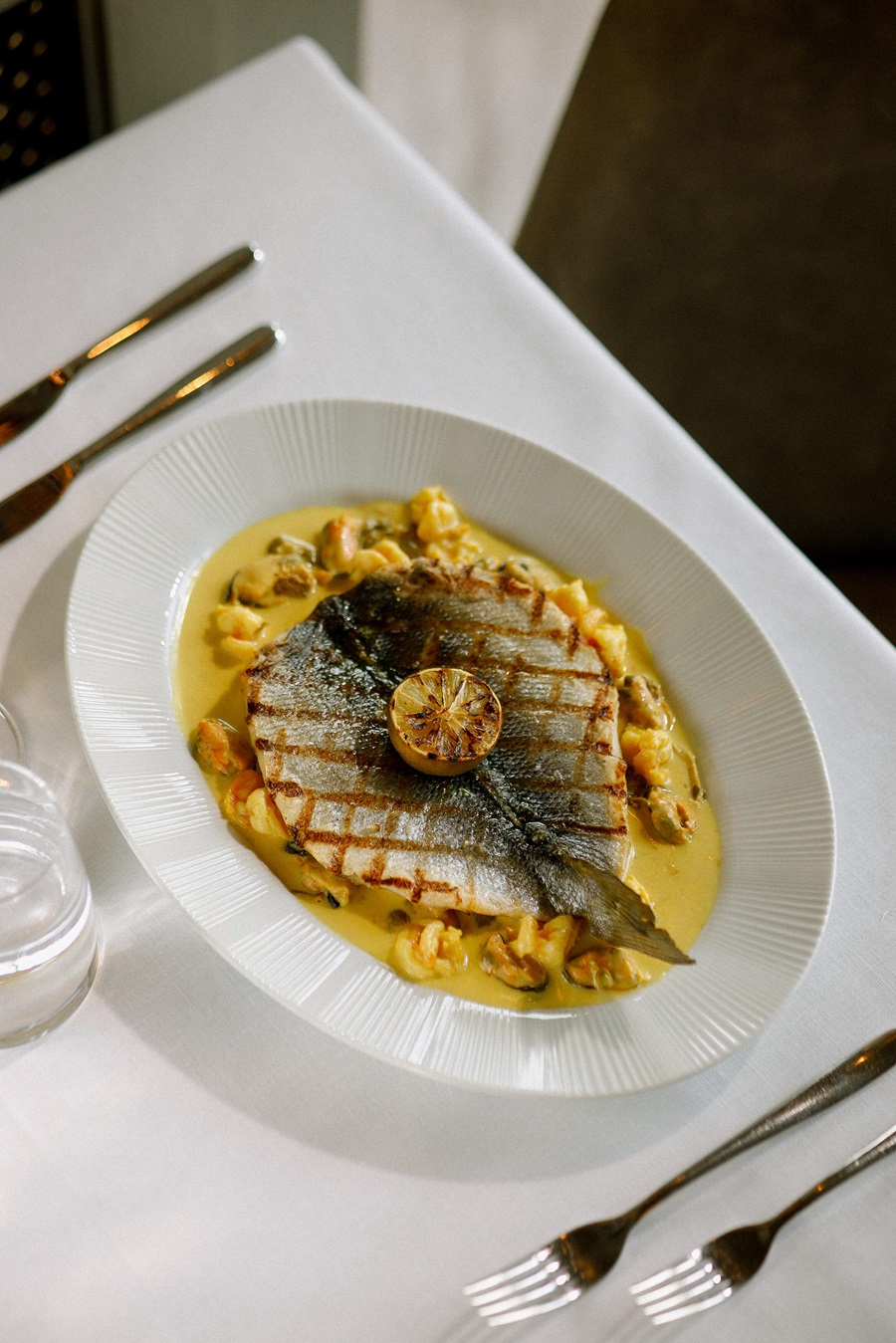Whole grilled fish on a bed of creamy yellow sauce with small potato dumplings, topped with a lemon half, plated on a white dish.