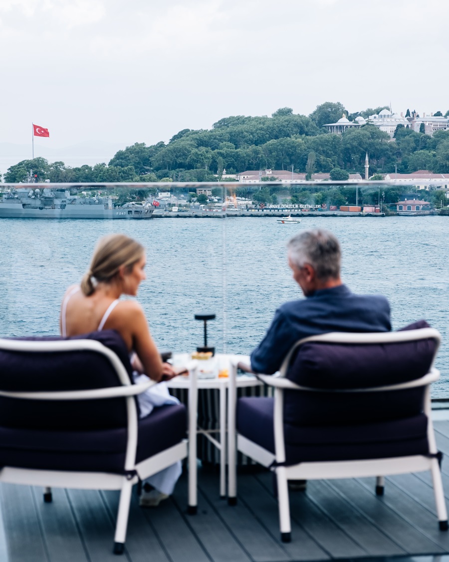 Couple dining on a terrace overlooking the water with ships and a hillside town in the background, Turkish flag visible across the bay.