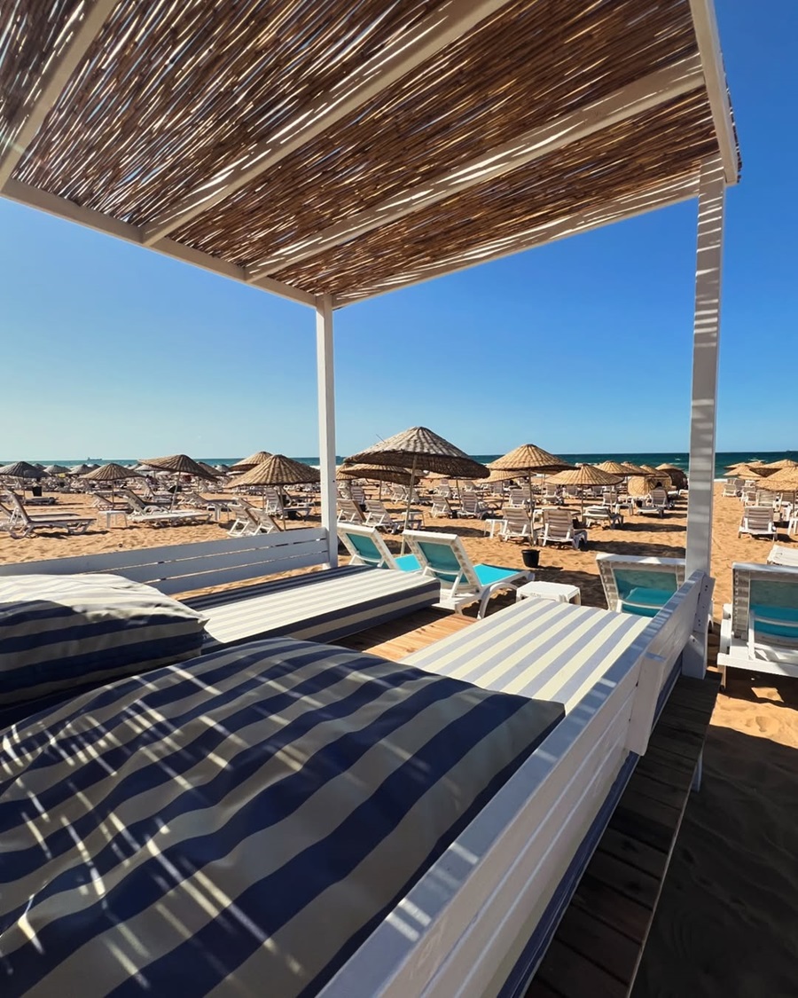 Beach scene with white sun loungers under a shaded pergola, rows of straw umbrellas, and blue sea in the distance.