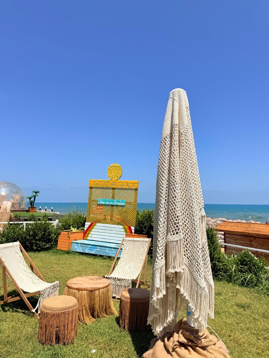 Beachside seating area with a large fringed macrame umbrella, woven chairs, and wooden stump stools overlooking the sea.