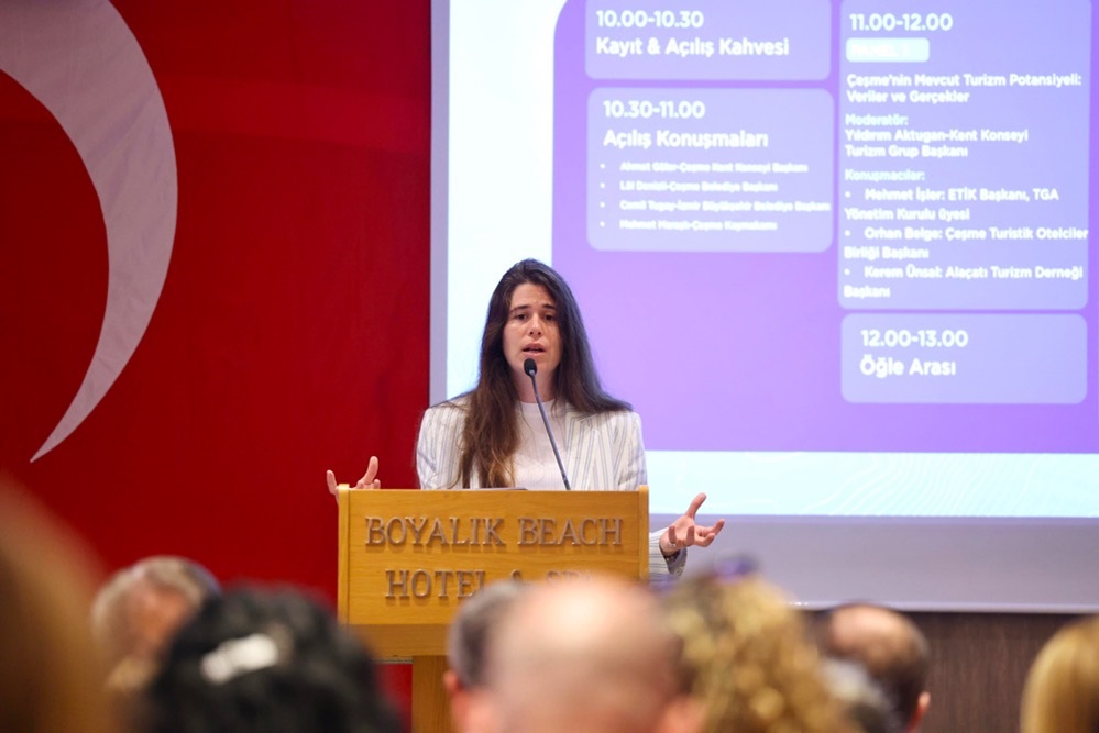 Woman speaking at a wooden podium in a hotel conference room, Turkish flag backdrop, and a projection screen behind her showing a schedule.