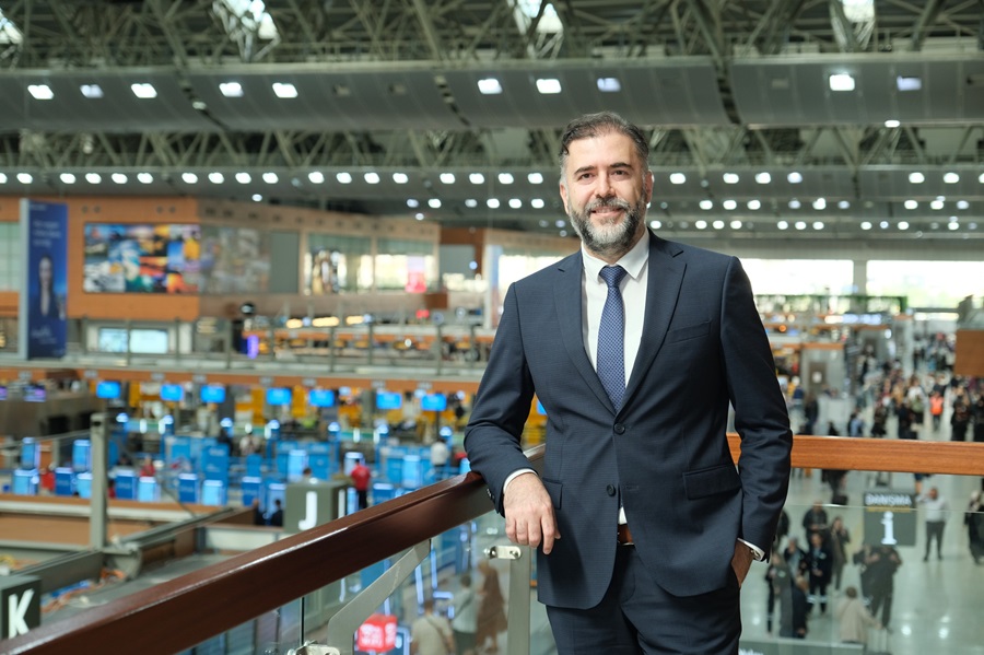 Smiling businessman in a navy suit leans on a balcony railing in a busy airport terminal beyond him shown with check-in counters and travelers.