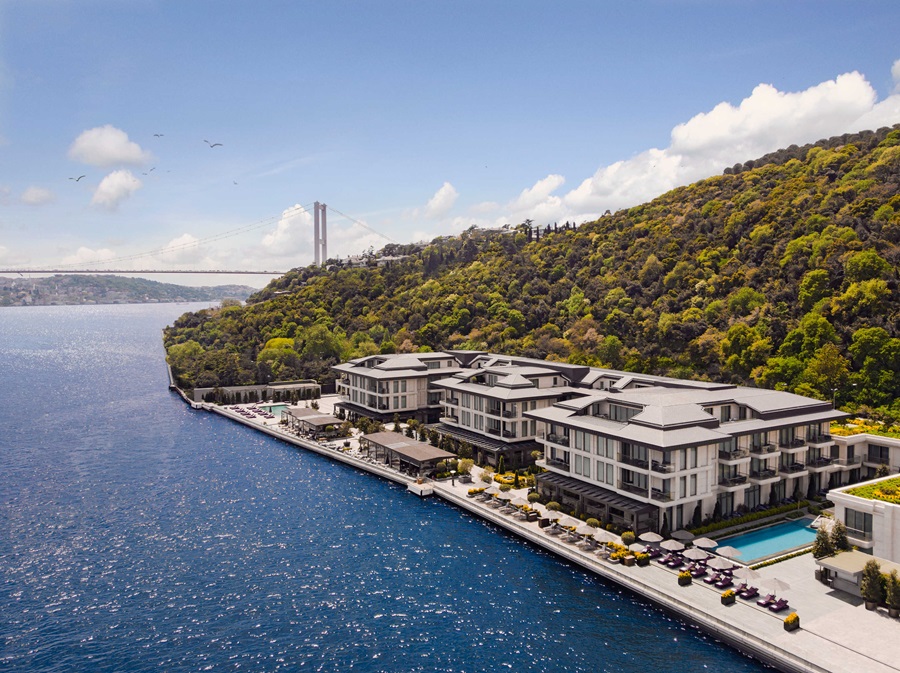 Modern waterfront resort with multi-story buildings along a blue bay and forested hills, with a distant bridge in the background.