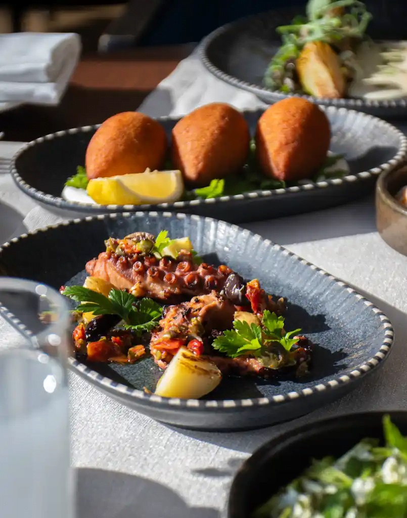 Plated octopus with peppers and herbs on a dark dish, lemon wedge nearby; croquettes on a separate plate in the background.