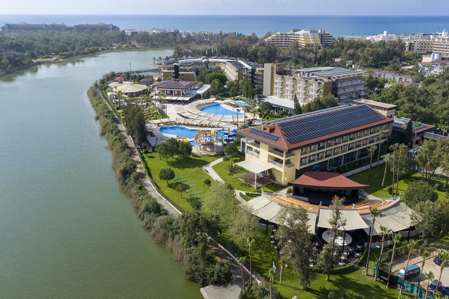 Aerial view of a seaside resort with multiple buildings, swimming pools, green lawns, and a canal along the left, with the ocean in the background.
