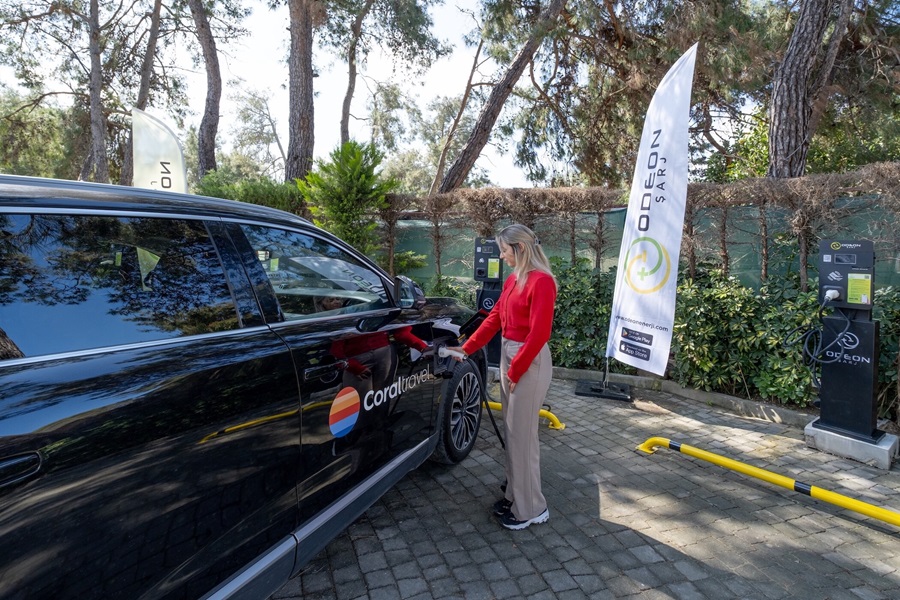 Woman in a red sweater plugs in a charger to a black electric van with a travel logo at an outdoor charging station.