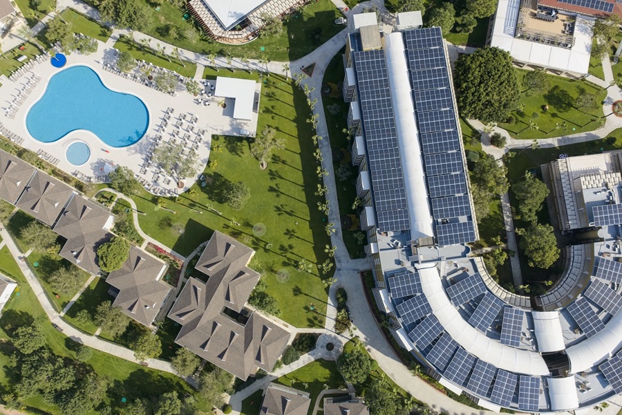 Aerial view of a resort complex with a kidney-shaped blue pool, sun loungers, and surrounding buildings with solar panels on some roofs.