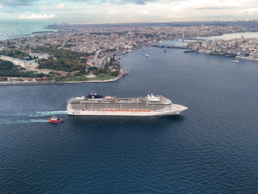 Large white MSC cruise ship sailing near a city coastline, with a red tugboat nearby.