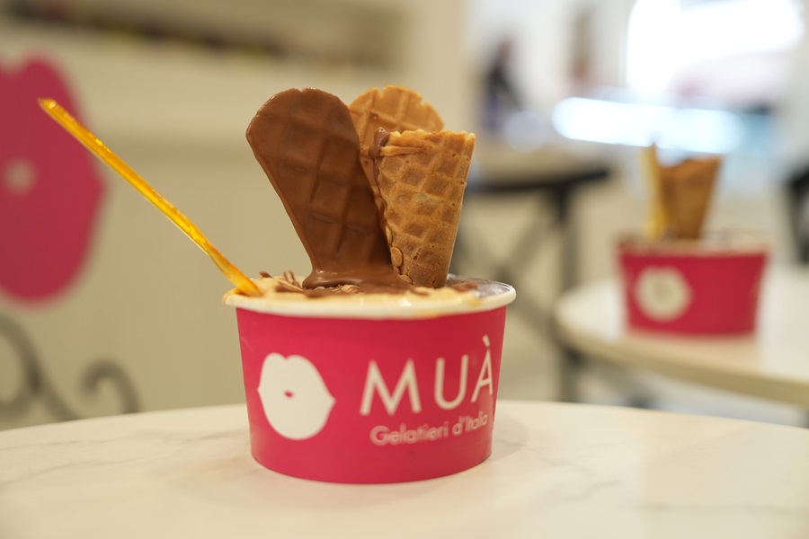 Pink MUÀ gelato cup with two chocolate-dipped waffle cones sticking out, on a marble table in a cafe.