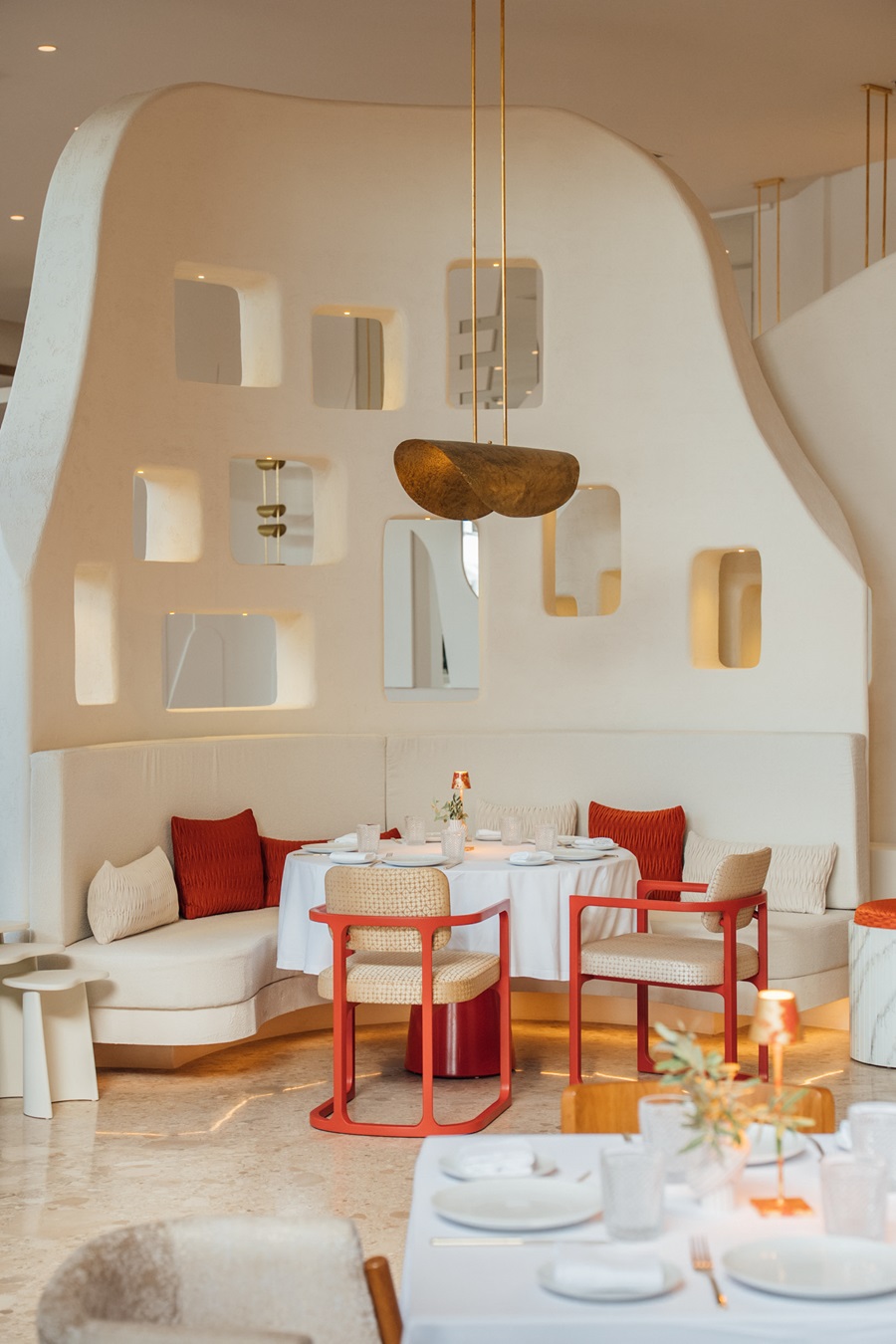 Curved white wall with square niches behind a round table set for dining in a modern restaurant, red chairs and white cushions nearby.