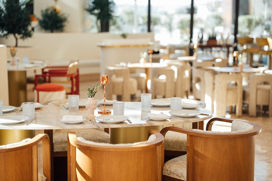 Round marble dining table set for six with white plates, glasses, and a small centerpiece in a bright restaurant interior with natural light.