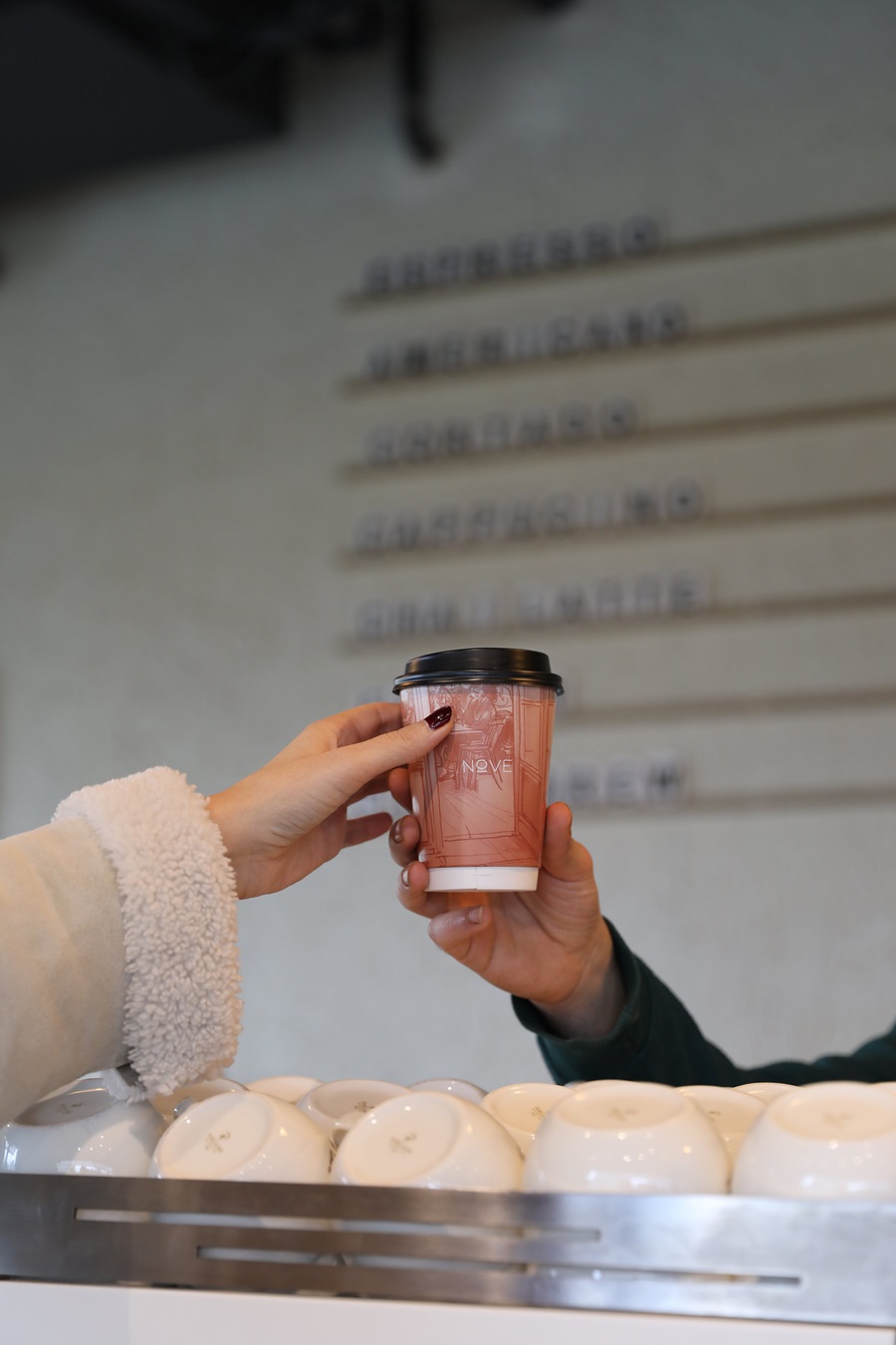 Two people exchange a pink take-away coffee cup with a black lid over a row of white cups at a cafe counter.