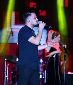 Male singer performing on stage with a microphone, backed by a female performer under bright stage lights.