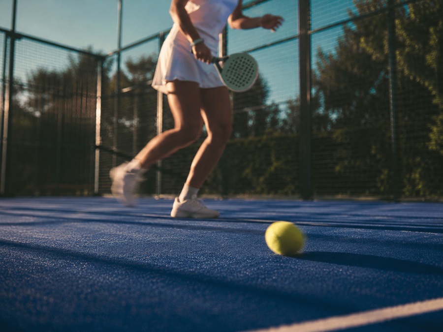 Tennis player in white on a blue outdoor court, racket mid-swing near a foreground tennis ball.