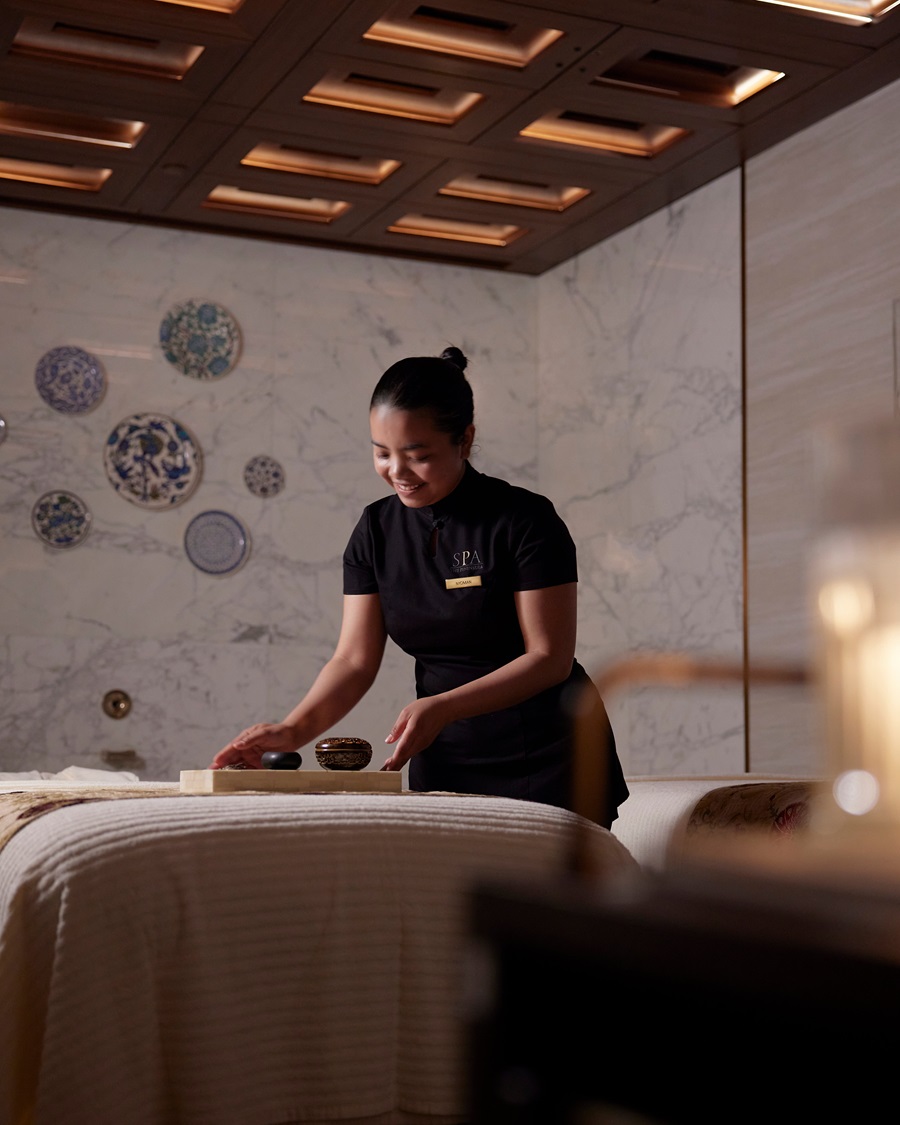 Spa therapist in a black uniform arranges stones on a tray beside a massage table in a marble-walled treatment room.
