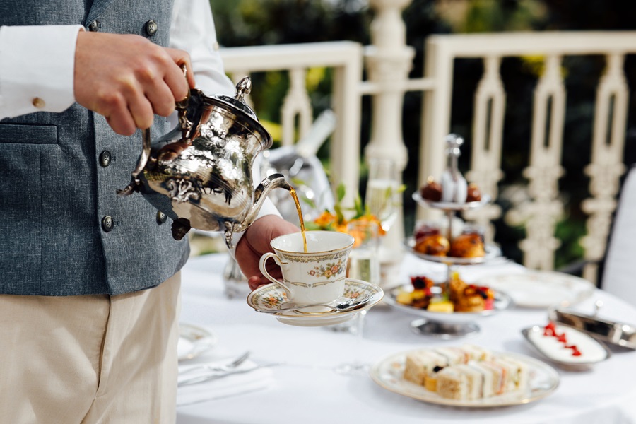 Person pours tea from a silver kettle into a floral teacup on a white-tablecloth outdoor tea spread.