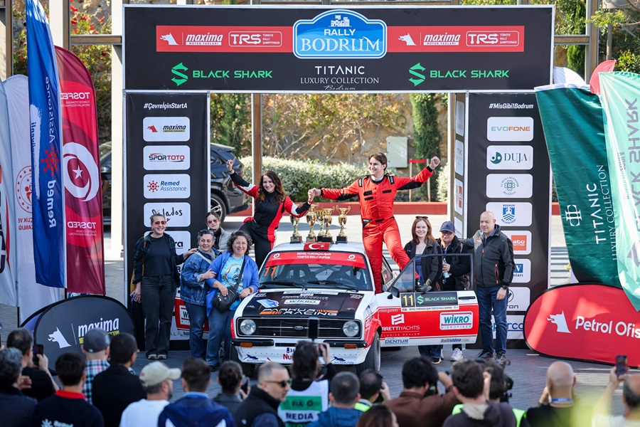 Rally Bodrum podium scene: a vintage rally car in front of a sponsor arch, two drivers in red suits stand on the car with raised arms and trophies on the roof, flanked by team members and a crowd of spectators beneath banners and flags.