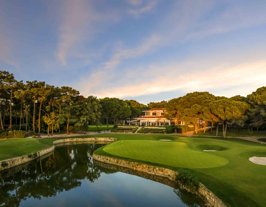 Golf course with a reflective water hazard, green, sand bunkers, and a clubhouse among trees at sunset.