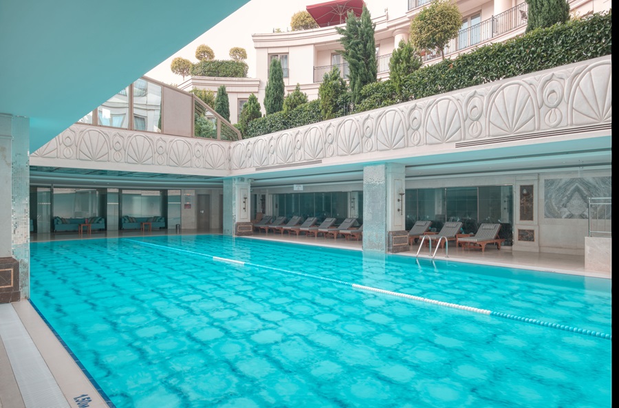 Indoor pool area with bright turquoise water, lounge chairs along the glass wall, and decorative balcony above.