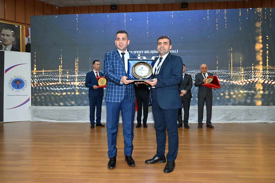 Two men in suits exchange a framed award on a stage, with others holding awards in the background.