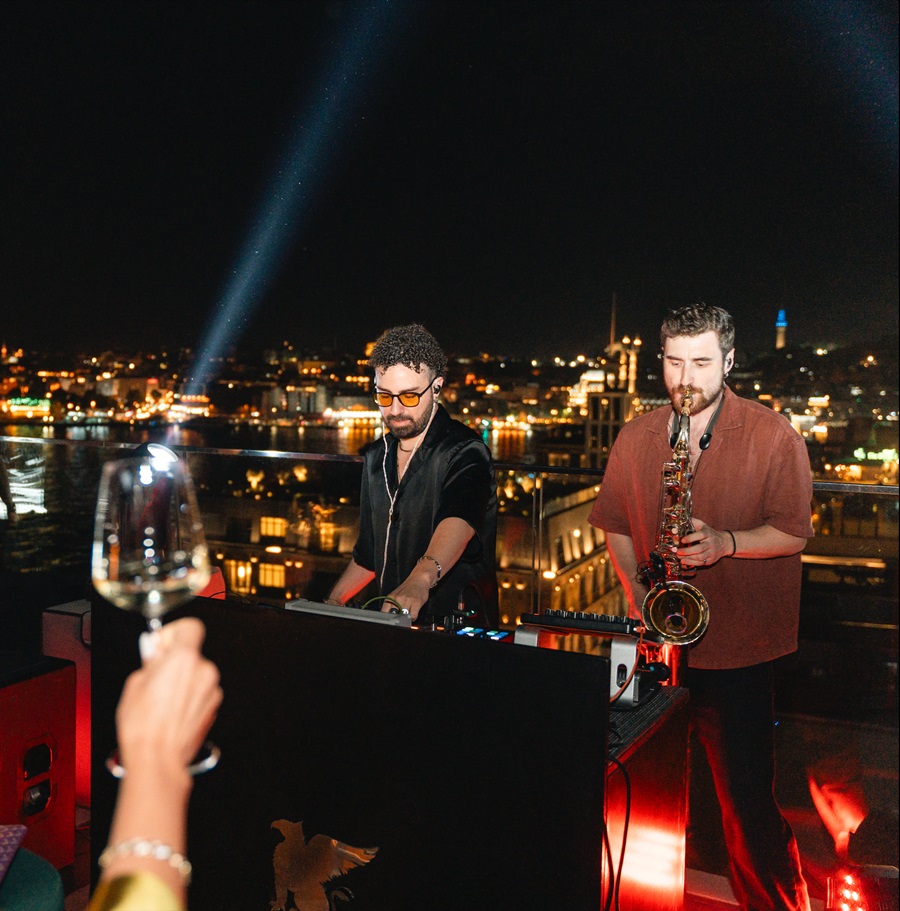 Two musicians perform on a rooftop at night: a DJ at a mixer and a saxophonist, with a glass of wine in the foreground and city lights in the background