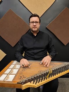 Man in a black shirt seated at a hammered dulcimer, playing with mallets.